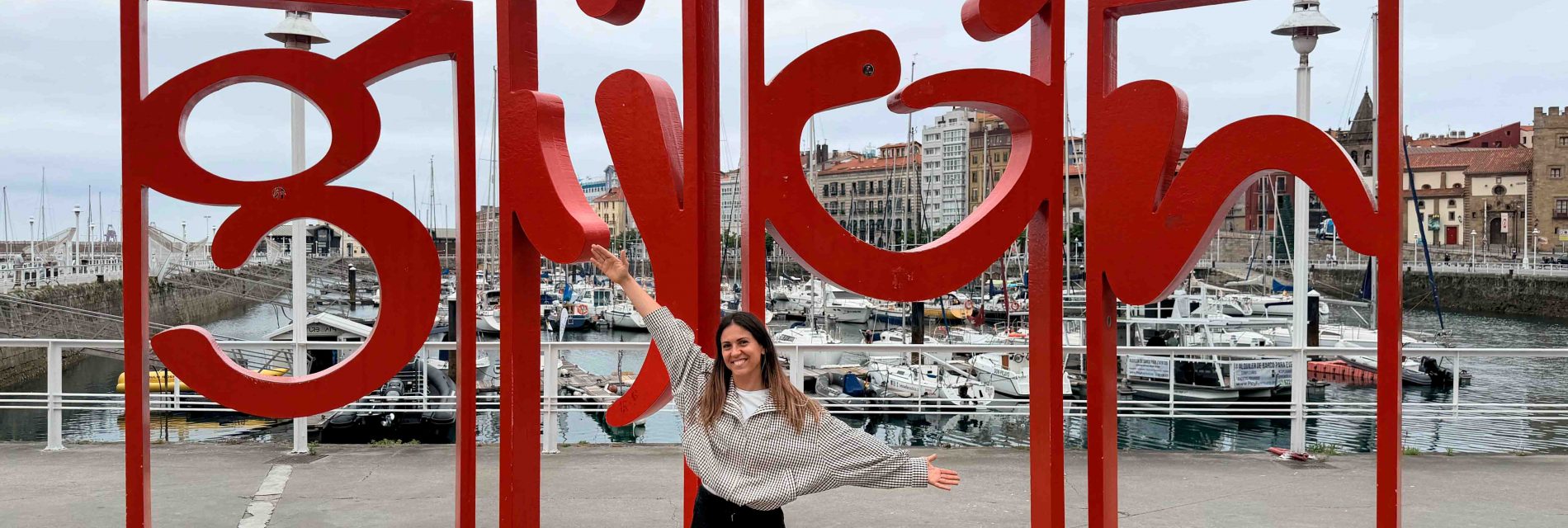 Gijon accesible: fotografía de mireia con los brazos abiertos delante de las letras miticas que pone Gijón