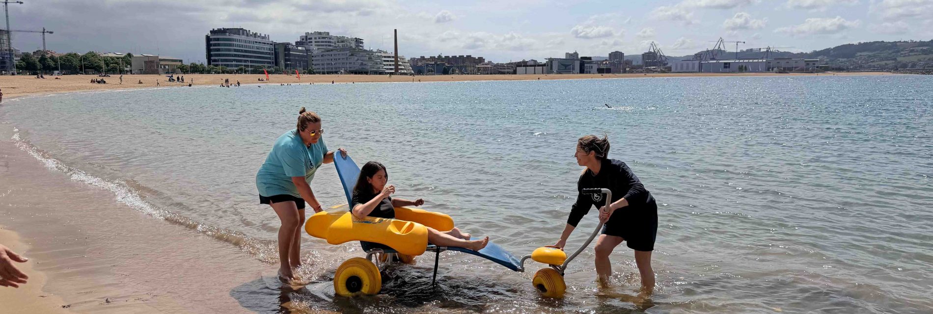 Actividades Accesibles en Gijón, La playa más accesible es poniente, mireia en una silla anfibia siendo trasladada al agua por el personal de la playa